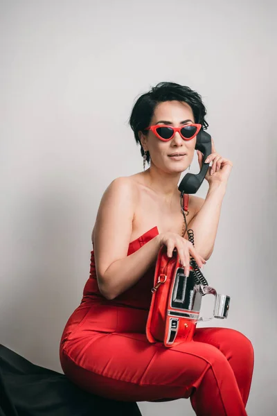 Stylish young lady in red dress talking on non-functional toy phone and holding her glamorous clutch in studio