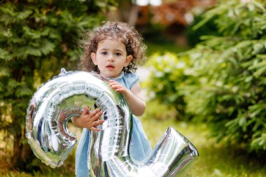 Doğum günü fotoşopu. Bahçede balon numarası tutan kıvırcık saçlı tatlı küçük kız kameraya dikkatlice bakıyor.