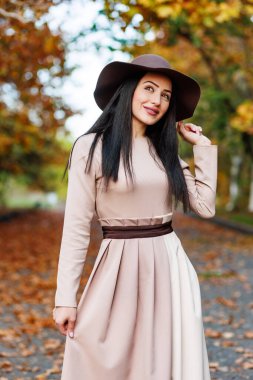 Parlak gözlü ve geniş kenarlı şapkalı tatlı genç kadın canlı sonbahar parkında kameraya poz veriyor..