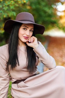 Şık ve modaya uygun bir kadın geniş kenarlı şapkası ve parlayan gözleriyle sonbahar parkında oturuyor..