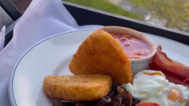 Patates kızartması, mantar, domates, sosis ve fırında fasulye içeren İngiliz kahvaltı tarifi, arka planda manzaralı kırsal bir manzara ile beyaz bir tabakta servis ediliyor. Yüksek kalite 4k görüntü