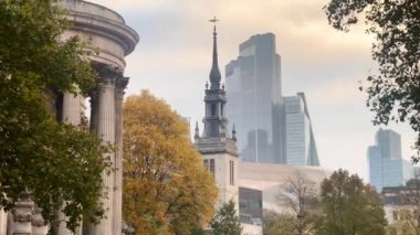 Londra 'da tarihi ve modern mimarinin sakin bir karışımı, sonbahar ağaçları ve arka planda çağdaş gökdelenler tarafından çerçevelenmiş bir kilise kulesi, yavaş çekim
