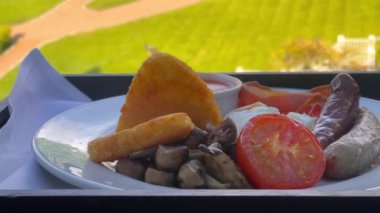 Patates kızartması, mantar, domates, sosis ve fırında fasulye içeren İngiliz kahvaltı tarifi, arka planda manzaralı kırsal bir manzara ile beyaz bir tabakta servis ediliyor. Yüksek kalite 4k görüntü