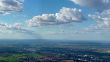 Uçsuz bucaksız açık araziler, mavi gökyüzünde dağınık bulutlar ve ufukta görünen uzak kasabalar, sakin ve geniş bir manzara yakalıyor.