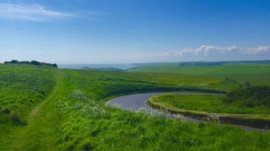 Uçsuz bucaksız mavi gökyüzünün altında dolambaçlı bir yol tarafından çerçevelenmiş yuvarlanan yeşil tepelerin ufuk çizgisiyle buluştuğu görkemli manzara. Bu huzurlu İngiliz kırsalında huzur ve doğal güzellik boldur.