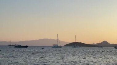 Gün batımının altın renkleri gökyüzünü huzurlu bir denizin üzerine boyar, uzaktaki yelkenli ve adalarla benekli, sakin ve rüya gibi bir deniz manzarası yaratır.