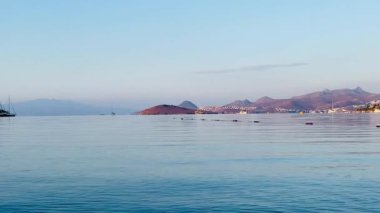 Sakin, cam gibi deniz ufukta uzak adalar ve yumuşak tepeler ile pastel bir gökyüzü altında sakin ve huzurlu bir kıyı manzarası oluşturur.