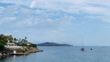 Türkiye 'nin Bodrum, Mugla kentindeki kıyı manzarasında yemyeşil, büyüleyici evleri olan kayalık bir kıyı şeridi ve yumuşak bir gökyüzü altındaki uzak adalara uzanan sakin mavi sular yer alıyor.