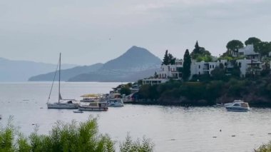 Sahil manzarası, ağaçlarla çevrili bir kıyıya demirlemiş yelkenliler, resimli beyaz evler ve sakin sulara karışan uzak dağlar.