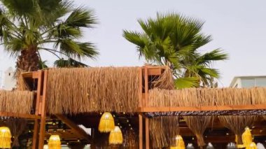 Sahil kenarındaki açık hava restoranı. Saf ahşap kirişler, saman çatısı ve sıcak ışıklandırma. Temiz bir gökyüzünün altında yemyeşil palmiye ağaçları ile çevrili.