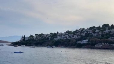 Türkiye 'nin Bodrum, Mugla kentindeki kıyı manzarasında yemyeşil, büyüleyici evleri olan kayalık bir kıyı şeridi ve yumuşak bir gökyüzü altındaki uzak adalara uzanan sakin mavi sular yer alıyor.