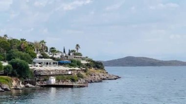 Türkiye 'nin Bodrum, Mugla kentindeki kıyı manzarasında yemyeşil, büyüleyici evleri olan kayalık bir kıyı şeridi ve yumuşak bir gökyüzü altındaki uzak adalara uzanan sakin mavi sular yer alıyor.