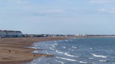 İngiltere, Doğu Sussex 'te Eastbourne, kumlu sahil İngiliz denizinin dalgalarıyla buluşuyor, berrak, sakin bir gökyüzü altında deniz kenarındaki mimarinin uzak bir görüntüsüyle,