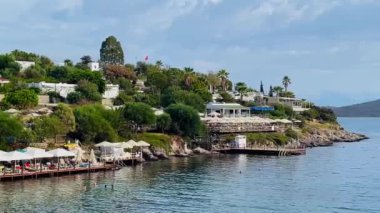 Türkiye 'nin Bodrum, Mugla kentindeki kıyı manzarasında yemyeşil, büyüleyici evleri olan kayalık bir kıyı şeridi ve yumuşak bir gökyüzü altındaki uzak adalara uzanan sakin mavi sular yer alıyor.