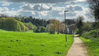 Huzurlu kırsal patika, yemyeşil çayırların arasında dolanıyor, gökyüzünün altında, tüylü beyaz bulutlarla kaplı, uzaktaki büyüleyici bir İngiliz köyüne doğru gidiyor.