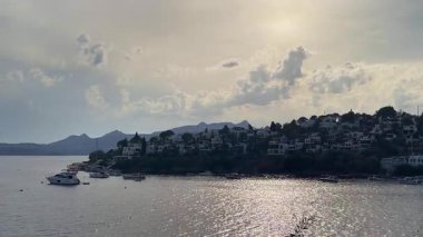 Türkiye 'nin Bodrum, Mugla kentindeki kıyı manzarasında yemyeşil, büyüleyici evleri olan kayalık bir kıyı şeridi ve yumuşak bir gökyüzü altındaki uzak adalara uzanan sakin mavi sular yer alıyor.