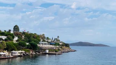 Türkiye 'nin Bodrum, Mugla kentindeki kıyı manzarasında yemyeşil, büyüleyici evleri olan kayalık bir kıyı şeridi ve yumuşak bir gökyüzü altındaki uzak adalara uzanan sakin mavi sular yer alıyor.