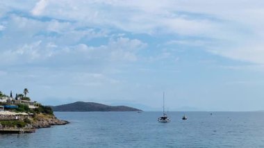 Sahil manzarası, ağaçlarla çevrili bir kıyıya demirlemiş yelkenliler, resimli beyaz evler ve sakin sulara karışan uzak dağlar.