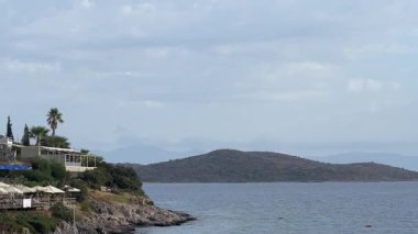 Türkiye 'nin Bodrum, Mugla kentindeki kıyı manzarasında yemyeşil, büyüleyici evleri olan kayalık bir kıyı şeridi ve yumuşak bir gökyüzü altındaki uzak adalara uzanan sakin mavi sular yer alıyor.