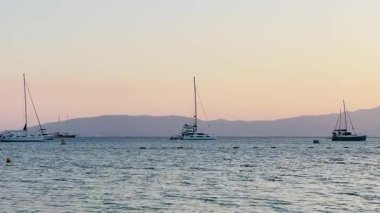 Gün batımının altın renkleri gökyüzünü huzurlu bir denizin üzerine boyar, uzaktaki yelkenli ve adalarla benekli, sakin ve rüya gibi bir deniz manzarası yaratır.