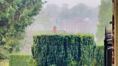 Kırsal İngiltere 'nin barışçıl ve atmosferik cazibesini çağrıştıran, yemyeşil ağaçlar ve çitlerle çerçevelenmiş, kısmen sisli, kırmızı tuğlalı bir binanın bulunduğu puslu kırsal bölge manzarası