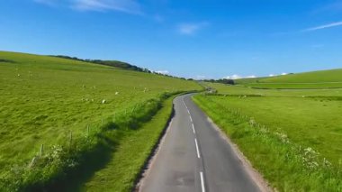 İngiliz kırsal yolu parlak mavi bir gökyüzünün altında canlı yeşil tepeler boyunca uzanır. Huzurlu manzara, kırsal sükunetin ve zamansız doğal güzelliğin cazibesini somutlaştırıyor.
