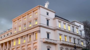 Londra 'da büyük bir neoklasik bina alacakaranlıkta sıcak bir şekilde parlıyor kara kara düşünen gökyüzünün altında, pencereleri içeriden hafifçe aydınlatılıyor.