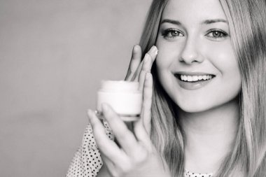 Gülümseyen sarışın kadın nemlendirici sürüyor, yüz kremi sürüyor, cilt kremi sürüyor. Klasik zarif monokrom portresi yumuşak ifade ve doğal güzellik konsepti.