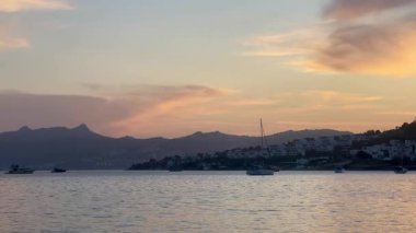 Altın akşam ışığı, Türkiye 'nin Bodrum kentindeki liman ve yamaç kenarındaki evlerin üzerine hafifçe düşüyor. Parlayan bulutların altında demirlemiş tekneler, ağır çekim.