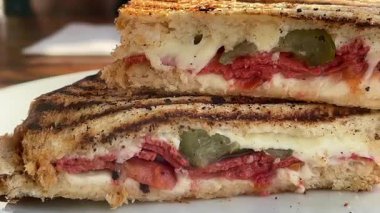 Eritilmiş peynir, domates ve biberle dolu altın ızgara sandviç deniz kenarındaki bir restoranın yanında, beyaz bir tabakta sıcak durur.