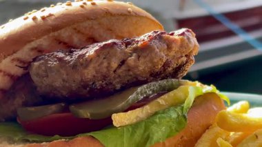 Marul, domates ve turşulu ızgara burger, deniz kenarındaki bir restoranda turkuaz bir tabakta altın cipsle servis edilir.