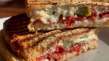 Eritilmiş peynir, domates ve biberle dolu altın ızgara sandviç deniz kenarındaki bir restoranın yanında, beyaz bir tabakta sıcak durur.