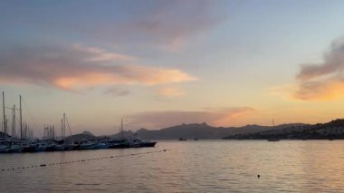 Altın akşam ışığı, Türkiye 'nin Bodrum kentindeki liman ve yamaç kenarındaki evlerin üzerine hafifçe düşüyor. Parlayan bulutların altında demirlemiş tekneler, ağır çekim.