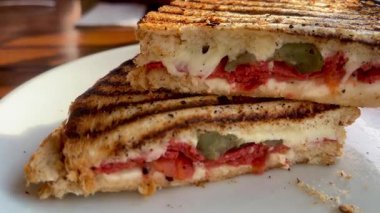 Eritilmiş peynir, domates ve biberle dolu altın ızgara sandviç deniz kenarındaki bir restoranın yanında, beyaz bir tabakta sıcak durur.