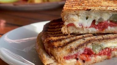 Eritilmiş peynir, domates ve biberle dolu altın ızgara sandviç deniz kenarındaki bir restoranın yanında, beyaz bir tabakta sıcak durur.