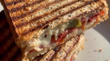 Eritilmiş peynir, domates ve biberle dolu altın ızgara sandviç deniz kenarındaki bir restoranın yanında, beyaz bir tabakta sıcak durur.