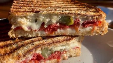 Eritilmiş peynir, domates ve biberle dolu altın ızgara sandviç deniz kenarındaki bir restoranın yanında, beyaz bir tabakta sıcak durur.