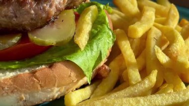 Marul, domates ve turşulu ızgara burger, deniz kenarındaki bir restoranda turkuaz bir tabakta altın cipsle servis edilir.