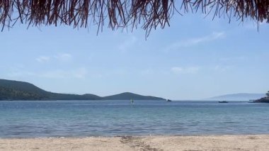 Sakin mavi sulara bakan sazlı bir şemsiyenin altından ve Türkiye 'nin Bodrum kenti yakınlarındaki kumlu sahilden yavaş çekim görüntüsü