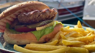 Marul, domates ve turşulu ızgara burger, deniz kenarındaki bir restoranda turkuaz bir tabakta altın cipsle servis edilir.