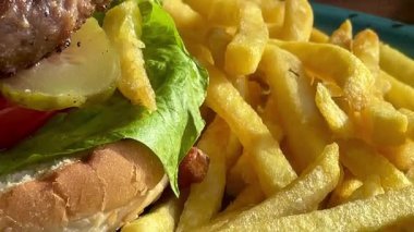 Marul, domates ve turşulu ızgara burger, deniz kenarındaki bir restoranda turkuaz bir tabakta altın cipsle servis edilir.