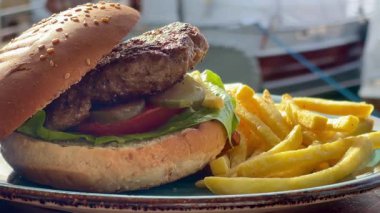 Marul, domates ve turşulu ızgara burger, deniz kenarındaki bir restoranda turkuaz bir tabakta altın cipsle servis edilir.