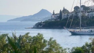 Bereketli kıyı yeşilliklerinde, Türkiye 'nin Bodrum kenti yakınlarındaki durgun mavi denizde bir yelkenli süzülüyor, tepe kenarındaki beyaz villalar ötesinde, ağır çekimde