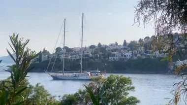 Bereketli kıyı yeşilliklerinde, Türkiye 'nin Bodrum kenti yakınlarındaki durgun mavi denizde bir yelkenli süzülüyor, tepe kenarındaki beyaz villalar ötesinde, ağır çekimde