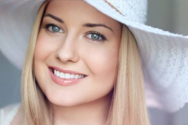 Yaz cilt bakımı ve beyaz dişler, kumsal şapkalı sarışın kadın yumuşak bir şekilde gülümsüyor, açık tenli ve açık renk makyajlı.