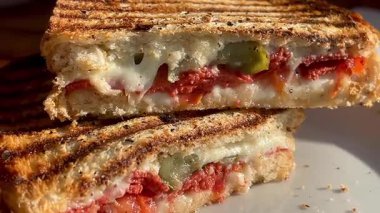 Eritilmiş peynir, domates ve biberle dolu altın ızgara sandviç deniz kenarındaki bir restoranın yanında, beyaz bir tabakta sıcak durur.