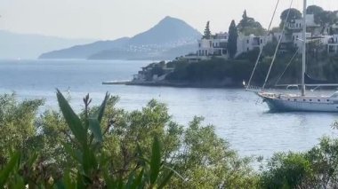 Bereketli kıyı yeşilliklerinde, Türkiye 'nin Bodrum kenti yakınlarındaki durgun mavi denizde bir yelkenli süzülüyor, tepe kenarındaki beyaz villalar ötesinde, ağır çekimde