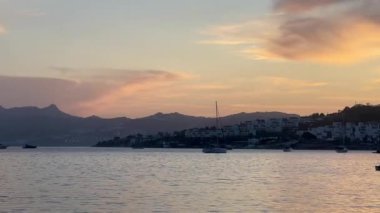 Altın akşam ışığı, Türkiye 'nin Bodrum kentindeki liman ve yamaç kenarındaki evlerin üzerine hafifçe düşüyor. Parlayan bulutların altında demirlemiş tekneler, ağır çekim.