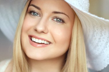 Yaz cilt bakımı ve beyaz dişler, kumsal şapkalı sarışın kadın yumuşak bir şekilde gülümsüyor, açık tenli ve açık renk makyajlı.