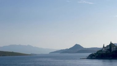 Mavi kıyı suları, Türkiye 'nin Bodrum kenti yakınlarındaki uzak dağlarla yarımadalar arasında uzanır ve sağ tarafta sakin bir sahil köyü bulunur.